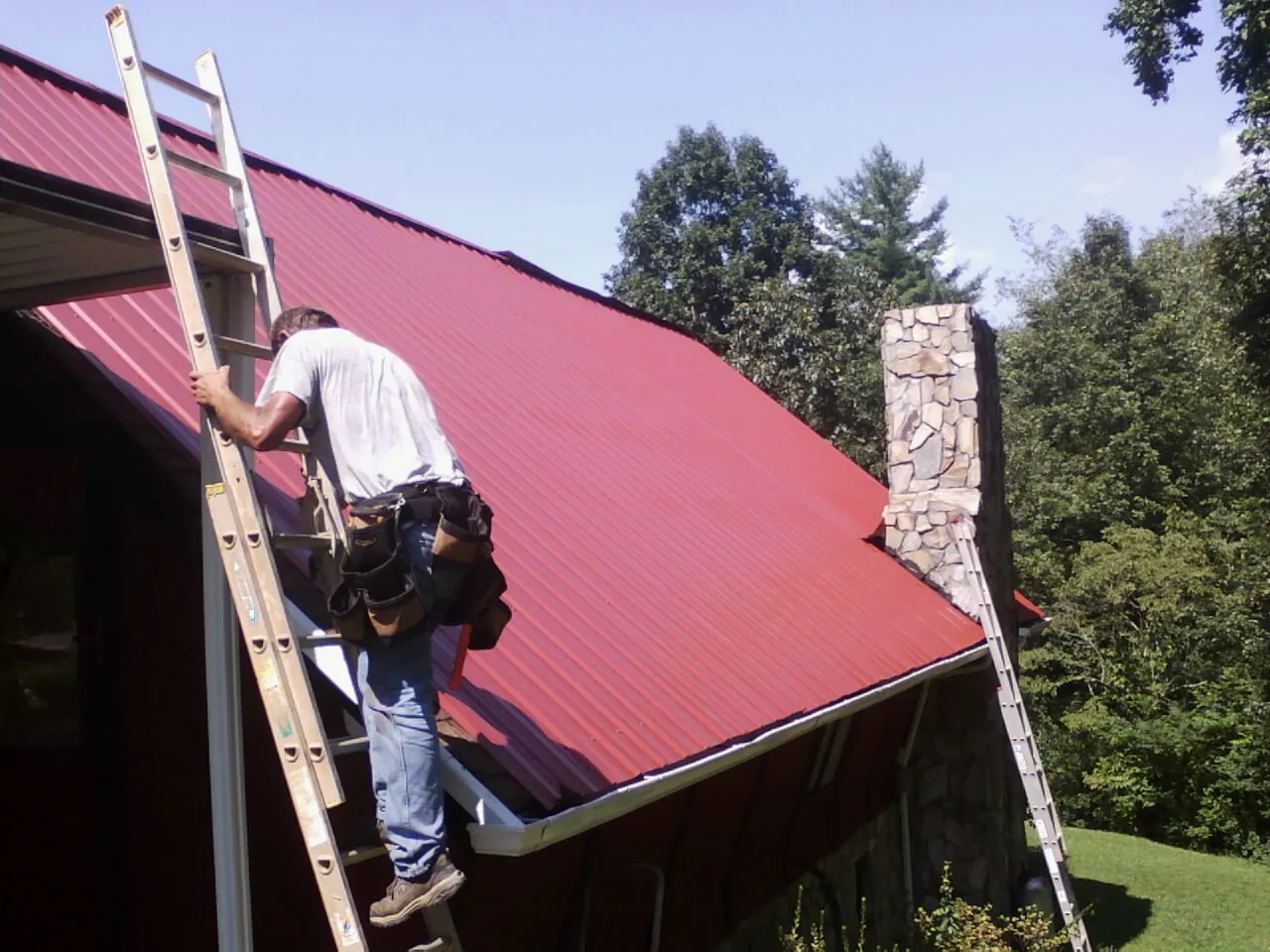 Roofer on a ladder beside a red metal roof and stone chimney — Soffit & Fascia Repair by Roofing Brothers in Mentone